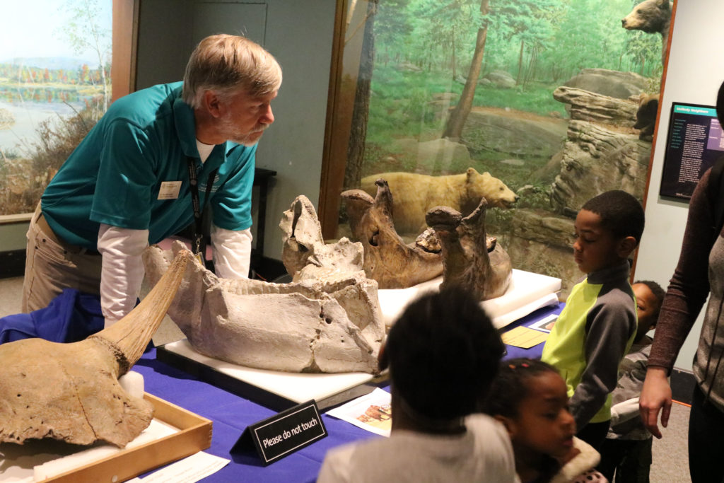 Kids under age 8 talk to male scientist about large fossils, such as a mastodon jaw, at center