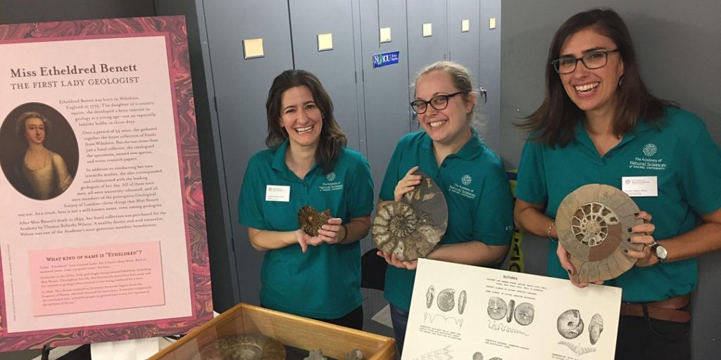 Three smiling women scientists holding fossils and standing in the Academy's Paleo collection storage lockers