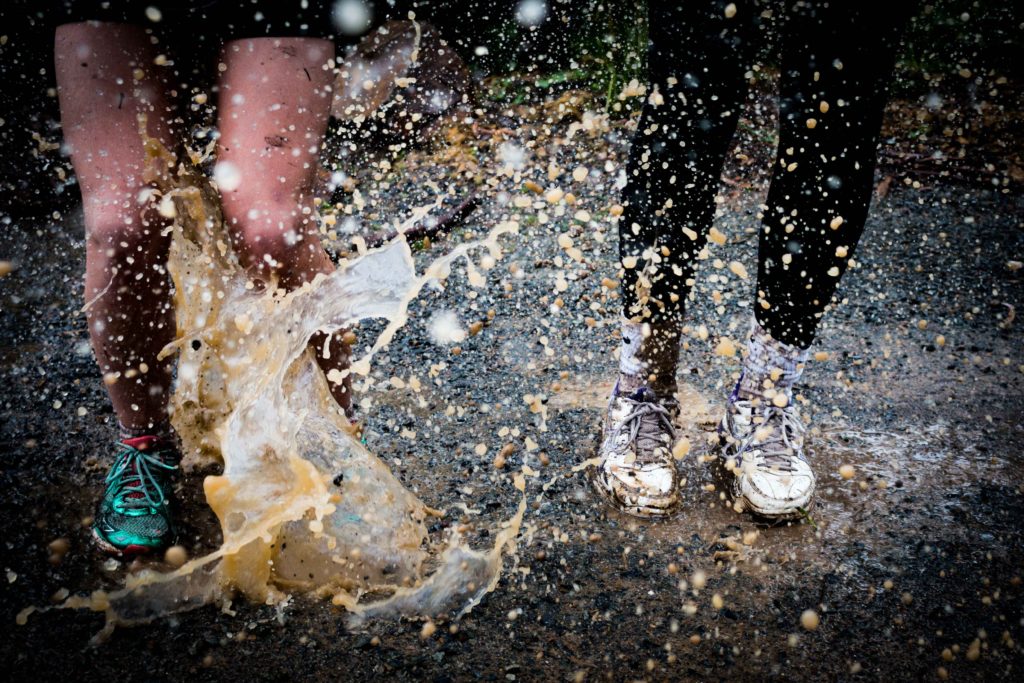 Two people splashing in muddy puddle. One wears shorts and teal sneakers, the other wears black leggings, white socks and white sneakers