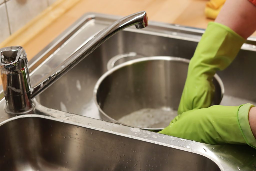 Person in neon green gloves washing large stainless steel pot in metal sink